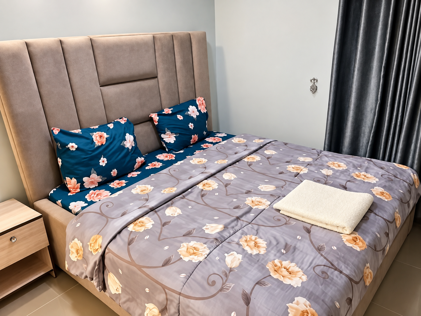 Room featuring padded beige headboard with floral-patterned bedding and bedside table